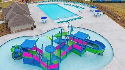 A colorful children's water play structure in a shallow pool with a large swimming pool, lounge chairs, and tennis courts nearby.